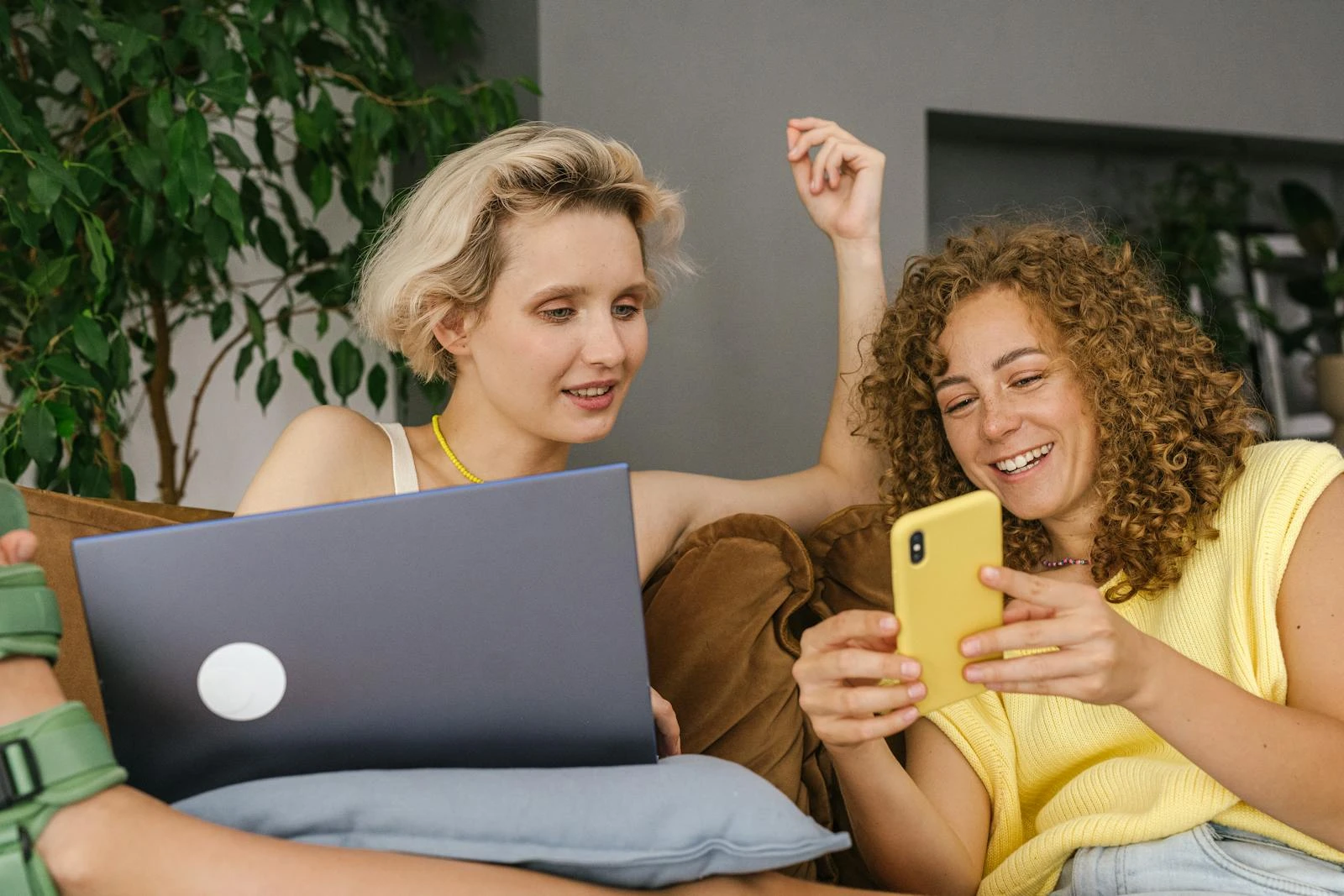 Two women sitting on a couch, engaging with a smartphone and laptop in a relaxed indoor setting.