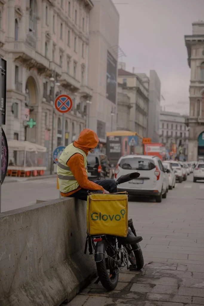Food delivery courier resting on a bicycle in a busy city street.