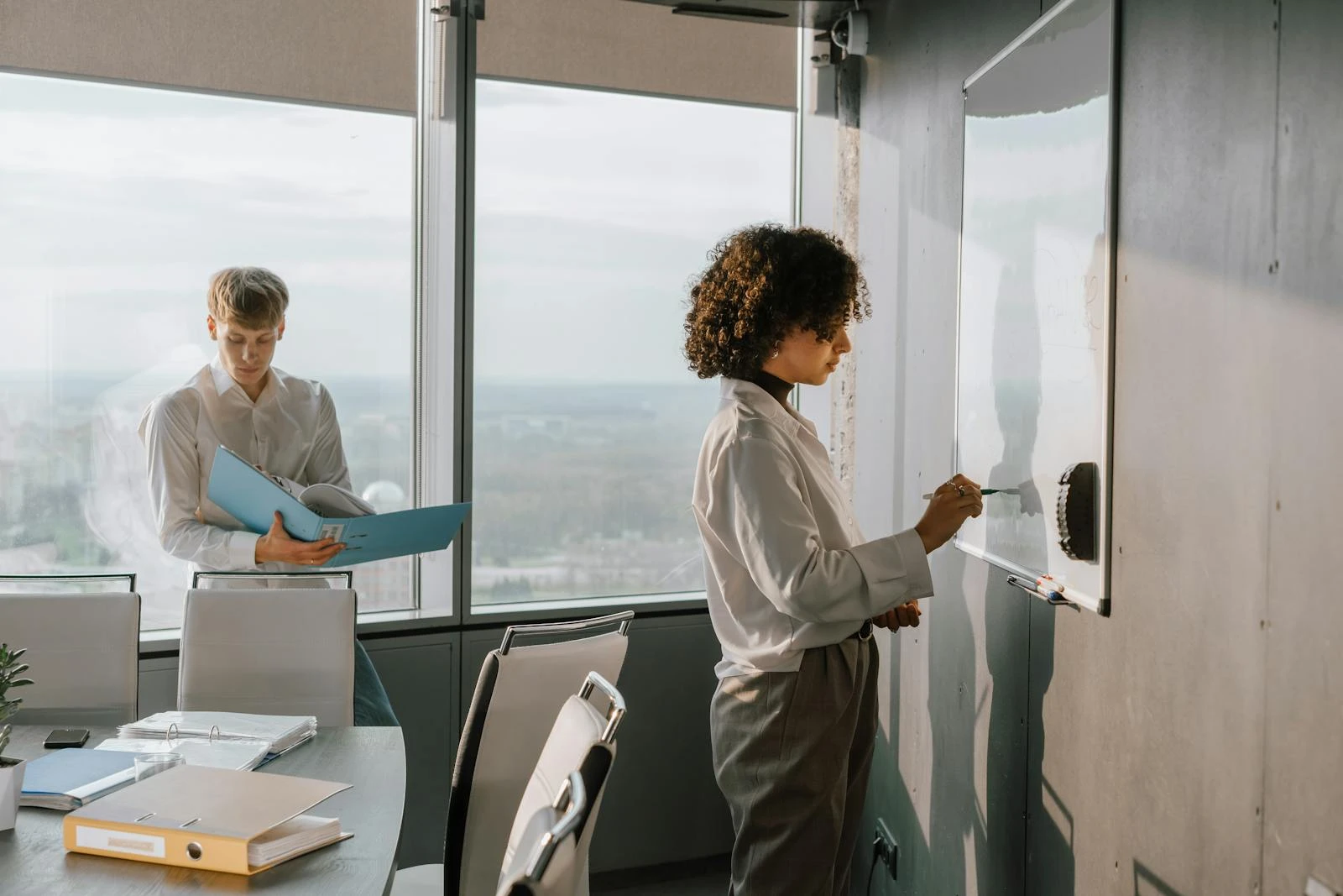 Two colleagues working in a modern office, one writes on a whiteboard while the other reviews documents.