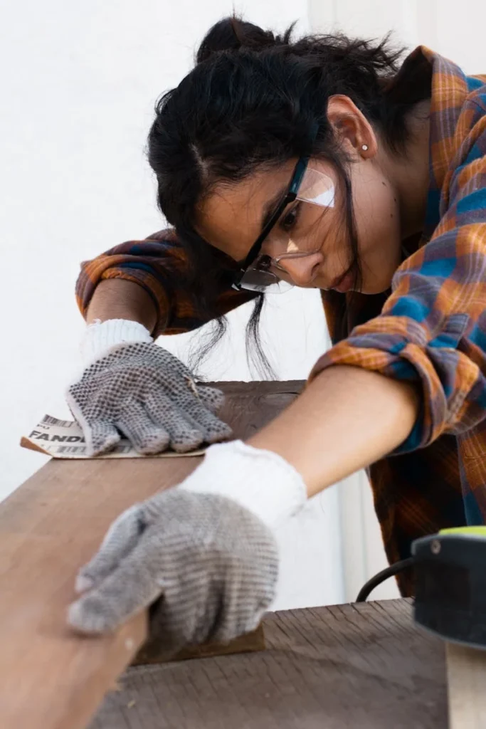 Focused woman wearing protective gear sands a wooden plank indoors.