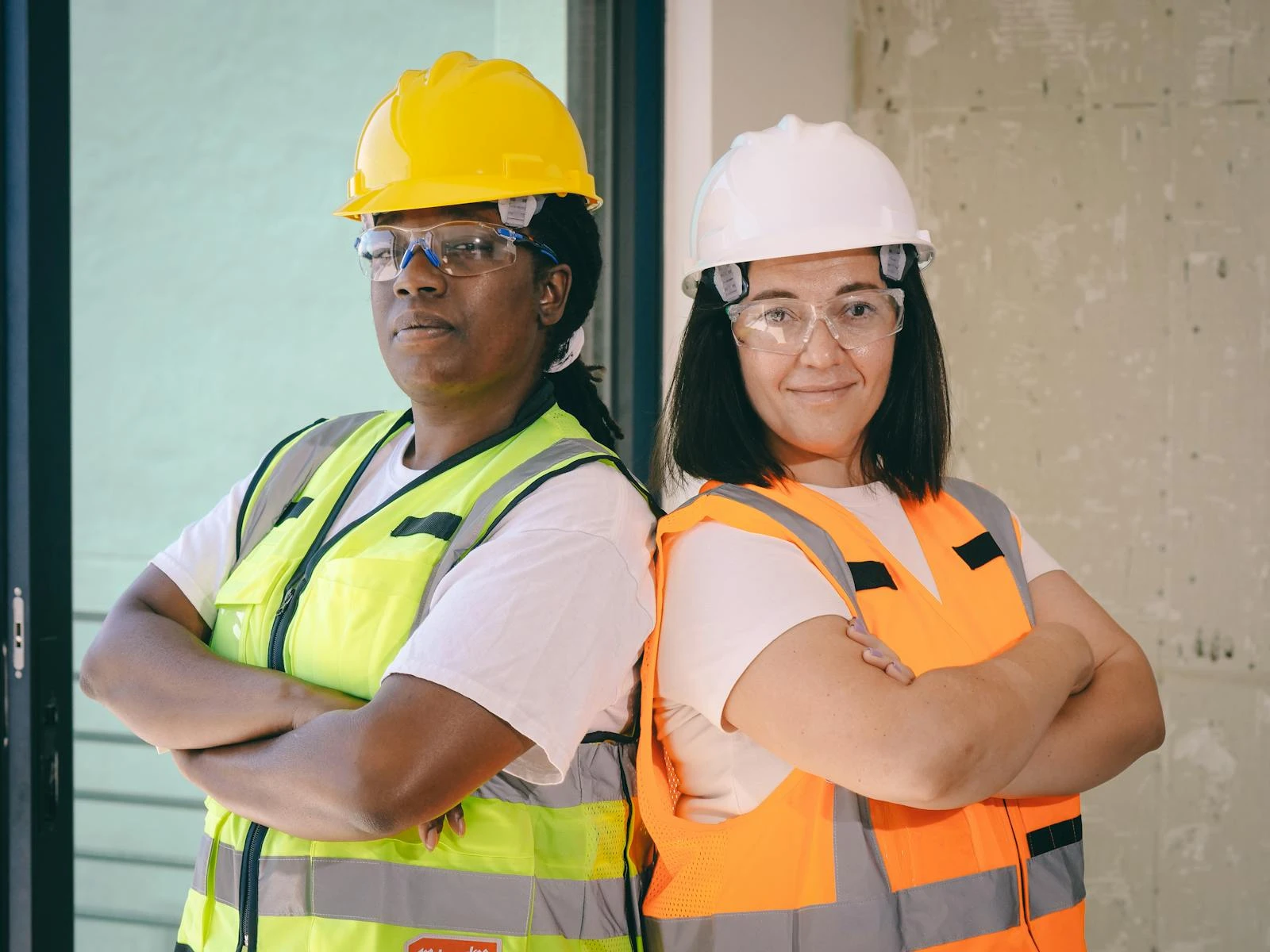 Two female engineers wearing safety gear stand confidently with crossed arms indoors.