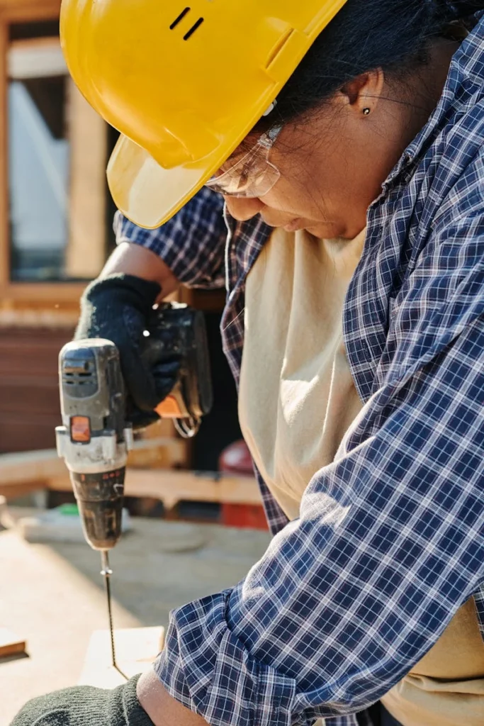 Woman in a hard hat using a power drill on wood indoors, showcasing focused craftsmanship and skill.