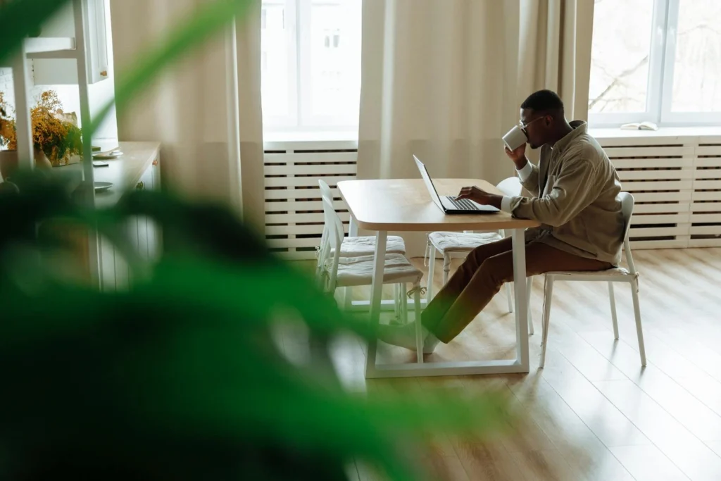 African American man working remotely from home using a laptop, enjoying coffee in a cozy setting.