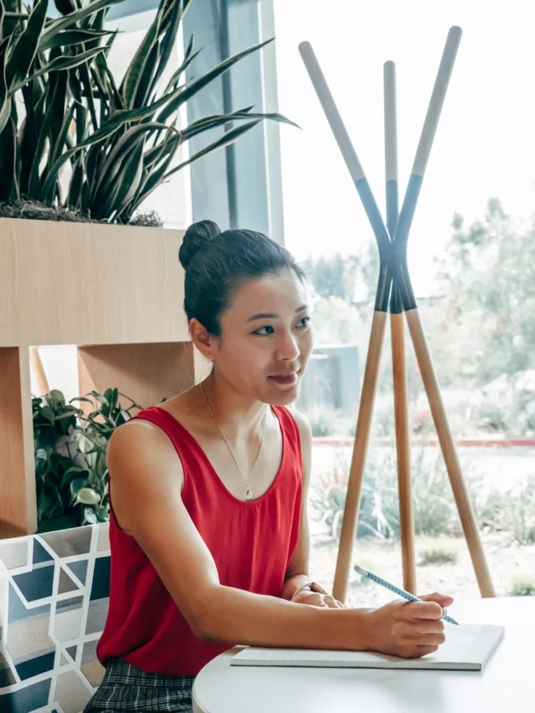Asian woman in a red tank top taking notes in a modern office setting, engaged in a planning session.